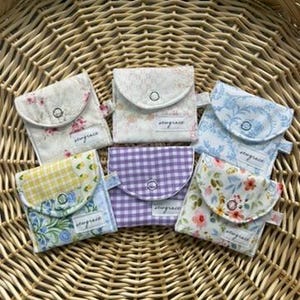 May include: A collection of small, fabric pouches with snap closures, each featuring a unique floral or patterned design. The pouches are in various colors, including white, blue, yellow, and purple, and are arranged on a woven basket.