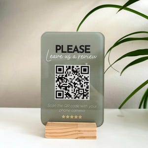 Könnte beinhalten: Ein rechteckiges Schild mit abgerundeten Ecken, das den Text "PLEASE leave us a review" und einen QR-Code zeigt. Es wird von einem kleinen Holzsockel getragen. Der Text "Scan the QR code with your phone camera" ist ebenfalls vorhanden.
