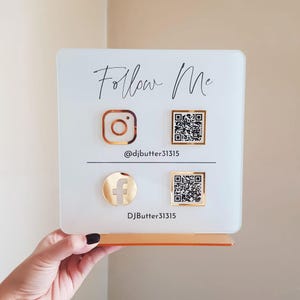 Peut inclure: Panneau en acrylique blanc avec l'inscription "Follow Me" en écriture élégante. Il présente des icônes Instagram et Facebook dorées, ainsi que des codes QR et le pseudonyme @djbutter31315. Le panneau est monté sur une base dorée.