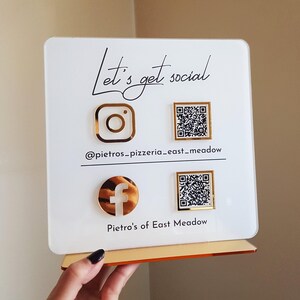 Op de afbeelding: Een wit acrylbord met gouden accenten, met social media-pictogrammen voor Instagram en Facebook, samen met QR-codes. Het bord leest "Let's get social" en bevat de bedrijfsnaam en locatie.