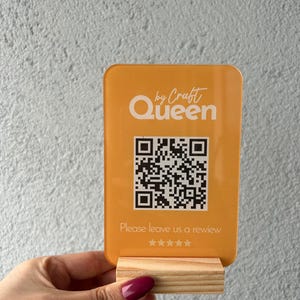 Op de afbeelding: Een oranje acrylbord met de tekst "by Craft Queen" en een QR-code. Het bord bevat ook de tekst "Please leave us a review" en sterrenbeoordelingen. Het bord wordt in een houten standaard gehouden.