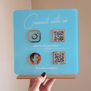 May include: A light blue acrylic sign with gold accents, featuring Instagram and Facebook logos, QR codes, and the text "Connect with us." The sign also displays the usernames @bakedbyani.maui and Bakedbyanimaui.