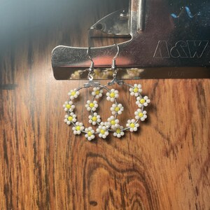 May include: Hoop earrings featuring daisy designs, crafted with white and yellow beads. The earrings hang from silver-toned hooks. The background is a wooden surface with a metal clip.