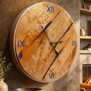 May include: A round wall clock featuring a natural light wood grain face with embedded blue resin accents. The clock has a silver-toned metal frame, black metal hands, and blue Roman numerals at the twelve, three, six, and nine o'clock positions.