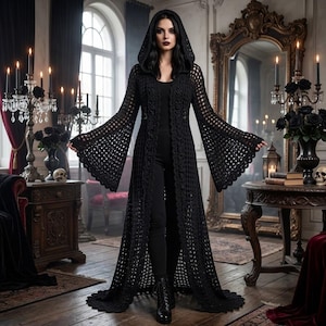 May include: A long, black, open-front crochet cardigan featuring a hood and wide, flared bell sleeves. The garment is floor-length with a scalloped hemline, worn over a black top and black trousers, paired with black lace-up boots.