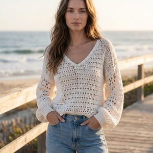 May include: A cream-colored long-sleeve sweater featuring an open-knit crochet pattern with a V-neckline and ribbed cuffs, paired with light blue denim jeans. The sweater has a loose, relaxed fit with slightly billowed sleeves and a textured design.