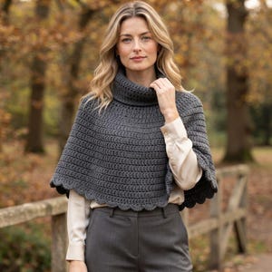 May include: A dark gray crocheted poncho featuring a high turtleneck collar and a scalloped hemline. The garment is worn over a light beige long-sleeved button-down shirt and dark gray trousers, showcasing a textured, hand-stitched pattern.