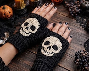 May include: A pair of black fingerless gloves featuring a crocheted white skull design on the back of each hand. The gloves have a ribbed cuff at the wrist and a textured knit pattern throughout, covering the palm and extending to the knuckles.