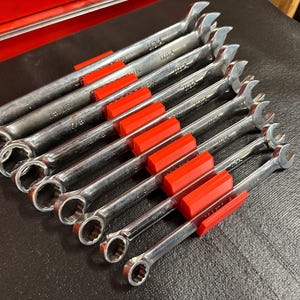May include: A set of eight silver wrenches with red holders, arranged diagonally on a dark surface. Each wrench is marked with "USA" and size measurements in inches. The wrenches are a combination of open-end and box-end styles.