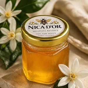 May include: A small glass jar of honey with a gold lid, labeled "NICA-D'OR MELIPONA RESERVE 100% RAW HONEY Origin: Nicaragua." The jar is surrounded by white flowers and green leaves. The honey is a golden amber color.