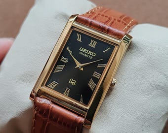 May include: A gold-toned Seiko Quartz watch with a rectangular black face and Roman numeral hour markers. The watch has a brown leather band with a crocodile pattern. The watch is displayed on a white pillow.