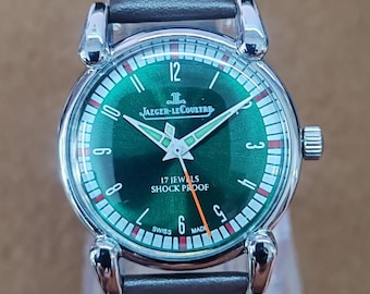 May include: A vintage Jaeger-LeCoultre wristwatch with a green dial and silver-tone hands and markers. The watch features a brown leather strap and the text "17 Jewels Shock Proof". The watch is a classic timepiece.