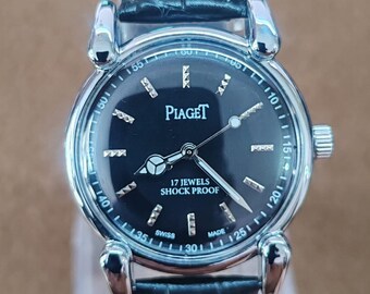 May include: A silver-toned Piaget wristwatch with a black face and a black leather band. The watch face features silver hands and markers, with the brand name and text "17 Jewels Shock Proof". The watch has a round face and a textured leather strap.