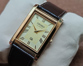 May include: A rectangular gold-toned Seiko Quartz watch with a cream-colored face and black leather band. The watch face features Roman numerals and a textured center. The watch is displayed on a white pillow.