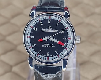 May include: A black Jaeger-LeCoultre wristwatch with a black leather band. The watch face is black with white and red accents, including the text "17 JEWELS SHOCK PROOF". The watch has a silver-colored case and a crown on the side.