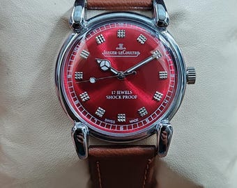 May include: A vintage Jaeger-LeCoultre wristwatch with a red dial and a brown leather strap. The watch features silver-tone hands and hour markers, and the text "17 JEWELS SHOCK PROOF" is visible on the dial. The watch is Swiss made.