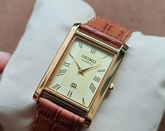 May include: A rectangular gold-toned Seiko Quartz watch with a cream-colored face and Roman numeral hour markers. The watch has a brown leather band with a crocodile pattern. The watch is displayed on a white pillow.