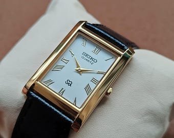 May include: A gold-toned rectangular Seiko Quartz watch with a white face and black leather band. The watch features Roman numeral hour markers and a small seconds dial. The watch is displayed on a cream-colored surface.