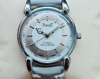 May include: A silver-toned Piaget watch with a light blue leather band. The watch face is silver with black hands and hour markers. The text "Piaget" is displayed above the center. The watch is marked "17 Jewels Shock Proof".