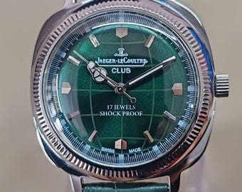 May include: A silver-toned Jaeger-LeCoultre Club watch with a green dial and a green leather strap. The watch face features the text "Jaeger-LeCoultre Club", "17 Jewels", and "Shock Proof". The watch is displayed on a clear stand.