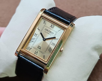 May include: A rectangular gold-toned Seiko Quartz watch with a black leather band. The watch face is a light gold color with black Roman numerals and hands. The watch is displayed on a white pillow.