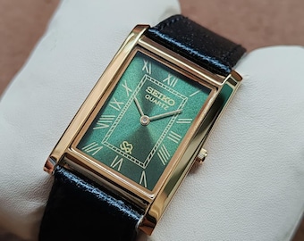 May include: A rectangular gold-toned Seiko Quartz watch with a green face and black leather band. The watch face features Roman numerals and the Seiko Quartz logo. The watch is displayed on a white pillow.