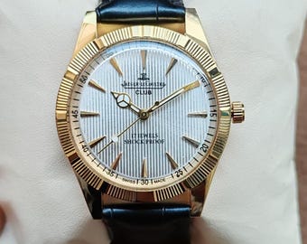 May include: A gold-toned wristwatch with a black leather band. The watch face is silver with vertical lines and the text "JAEGER-LECOULTRE CLUB 17 JEWELS SHOCK PROOF SWISS MADE". The watch is on a cream-colored pillow.