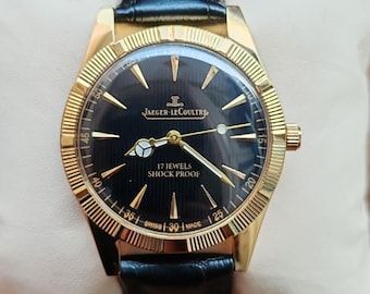 May include: A gold-toned Jaeger-LeCoultre wristwatch with a black dial and a black leather strap. The watch face features gold hands and markers, with the text "17 JEWELS SHOCK PROOF". The watch is displayed on a white surface.
