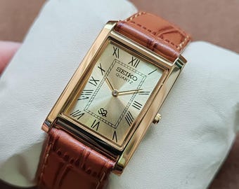 May include: A gold-toned Seiko Quartz watch with a rectangular face and Roman numeral hour markers. The watch has a brown leather band with a crocodile pattern. The watch is displayed on a white pillow.