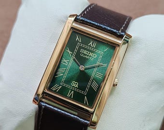 May include: A rectangular gold-toned Seiko Quartz watch with a dark green face and Roman numeral hour markers. The watch has a brown leather band. The watch is displayed on a white pillow.