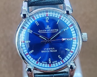 May include: A silver-toned Jaeger-LeCoultre watch with a blue dial and black leather strap. The watch face features white and red markers, and the text "17 JEWELS SHOCK PROOF". The watch has a black leather strap.