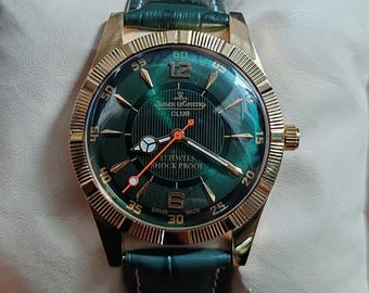 Può includere: Un orologio da polso dorato con quadrante verde e cinturino in pelle verde. Il quadrante dell'orologio presenta il testo "JAEGER LECOULTRE CLUB" e "17 JEWELS SHOCK PROOF". L'orologio ha una lunetta strutturata e una corona dorata.