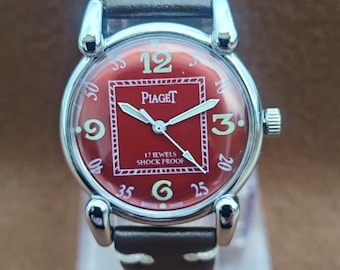 May include: A vintage Piaget wristwatch with a red square dial, featuring the brand name and the text "17 JEWELS SHOCK PROOF". The watch has a silver-tone case, a brown leather strap, and displays the numbers 12, 3, 6, 9, 25, 35, 40, and 50.