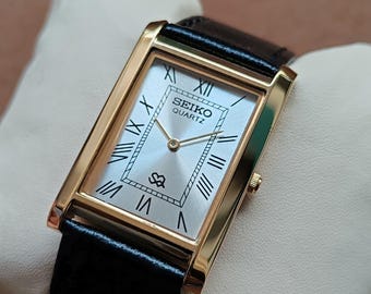 May include: A rectangular gold-toned Seiko Quartz watch with a silver face and black leather band. The watch face features Roman numerals and the Seiko logo. The watch is displayed on a white surface.