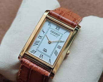 May include: A rectangular gold-toned Seiko Quartz watch with a white face and Roman numeral hour markers. The watch has a brown leather band with a crocodile pattern. The watch is displayed on a white pillow.