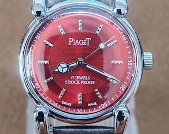 May include: A Piaget wristwatch with a red dial and silver-tone case. The watch features a black leather strap and the text "17 Jewels Shock Proof". The dial has silver markers and hands.