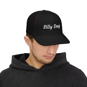 Op de afbeelding: Zwarte truckerpet met de tekst "Silly Dog" in witte letters. De pet heeft een gebogen klep en een mesh achterkant. De persoon draagt een zwarte hoodie.