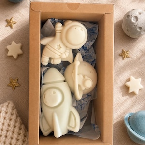 May include: A rectangular cardboard box containing three off-white shaped soaps: an astronaut, a rocket ship, and a planet with rings. The soaps are nestled in blue and white patterned tissue paper inside the box, which is open at the top.