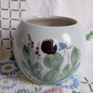 May include: A spherical ceramic planter with a smooth, light gray finish. The exterior features a hand-painted botanical design consisting of green leaves, small red berries, a single dark purple circular element, and a small blue flower.
