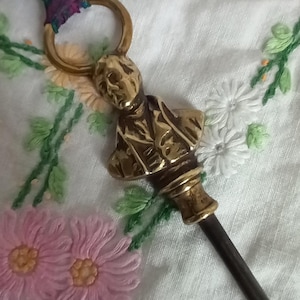 May include: A brass letter opener with a man's bust, attached to a dark handle and a ring. The opener has a decorative ribbon in purple and green. The background is a white cloth with embroidered flowers.