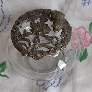 May include: A clear glass bowl with a decorative metal lid. The lid features a floral and animal design in a silver-gray color. The bowl is round and sits on a white surface with floral embroidery.