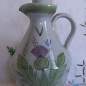 May include: A ceramic jug with a handle and a stopper. The jug is decorated with a purple thistle, green leaves, and other floral accents. The jug is light grey with a green rim.
