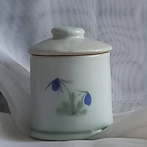 May include: A small, off-white ceramic jar with a lid. The jar is decorated with two bluebell flower illustrations. The lid has a small knob for easy removal. The jar is cylindrical and sits on a slightly raised base.
