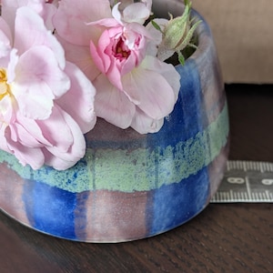 May include: A ceramic planter with a glazed finish in horizontal bands of blue, green, and brown, filled with light pink roses. A white measuring tape is positioned at the base, showing a width of approximately 3.5 inches across the vessel.