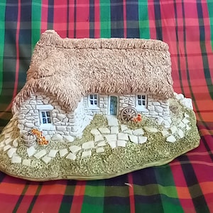 May include: A small, stone cottage with a thatched roof, a green door, and a wheelbarrow in front. The cottage is surrounded by a stone path and green grass.