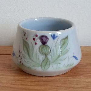 May include: A small, white ceramic bowl with a light blue interior. The exterior is decorated with a hand-painted design featuring a purple thistle, green leaves, red berries, and blue flowers. The bowl sits on a wooden surface.