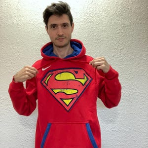 Puede incluir: Sudadera roja con interior azul y un gran logotipo de Superman en la parte delantera. El logotipo es amarillo con un contorno negro y una forma de diamante rojo. La sudadera tiene un bolsillo de canguro y un logotipo de Nike.