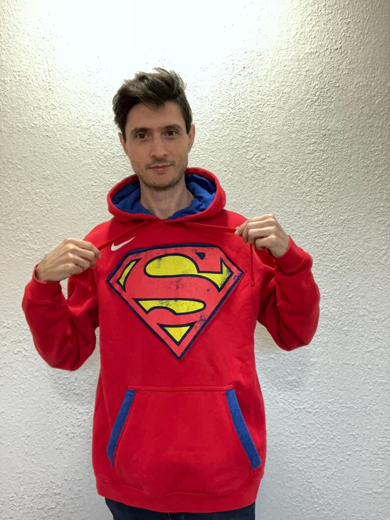 Puede incluir: Sudadera roja con interior azul y un gran logotipo de Superman en la parte delantera. El logotipo es amarillo con un contorno negro y una forma de diamante rojo. La sudadera tiene un bolsillo de canguro y un logotipo de Nike.