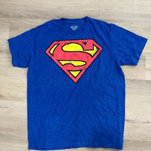 Puede incluir: Camiseta azul con el logo de Superman en rojo, amarillo y negro. El logo de Superman tiene forma de diamante con la letra "S" en el centro. La camiseta tiene mangas cortas y cuello redondo.