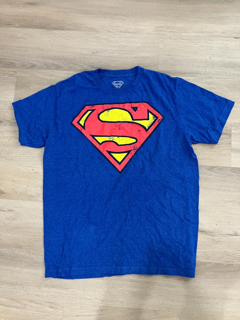 Puede incluir: Camiseta azul con el logo de Superman en rojo, amarillo y negro. El logo de Superman tiene forma de diamante con la letra "S" en el centro. La camiseta tiene mangas cortas y cuello redondo.